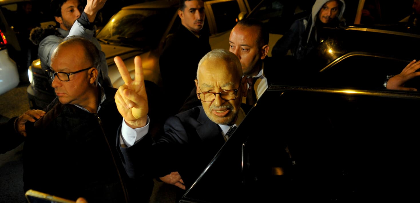An elderly man in a suit and glasses steps out of a black vehicle on a crowded nighttime street, with bodyguards around him, flashing the two fingered 