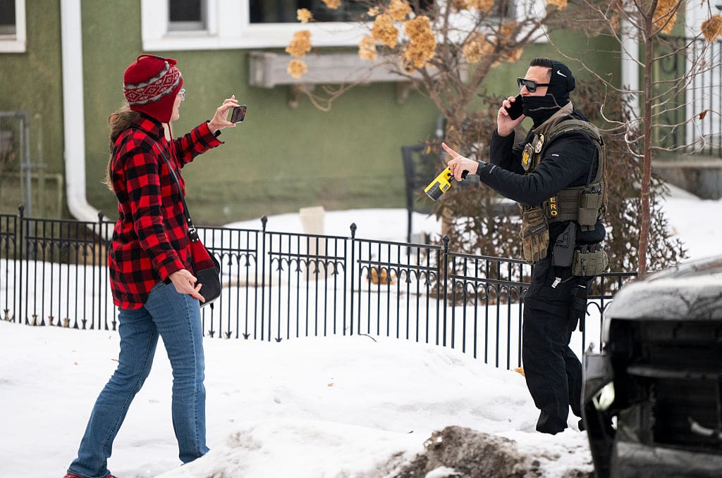 An ICE agent holding a taser points at a woman holding a phone after one of their vehicles got a flat tire on Penn Avenue on February 5, 2026 in Minneapolis, Minnesota