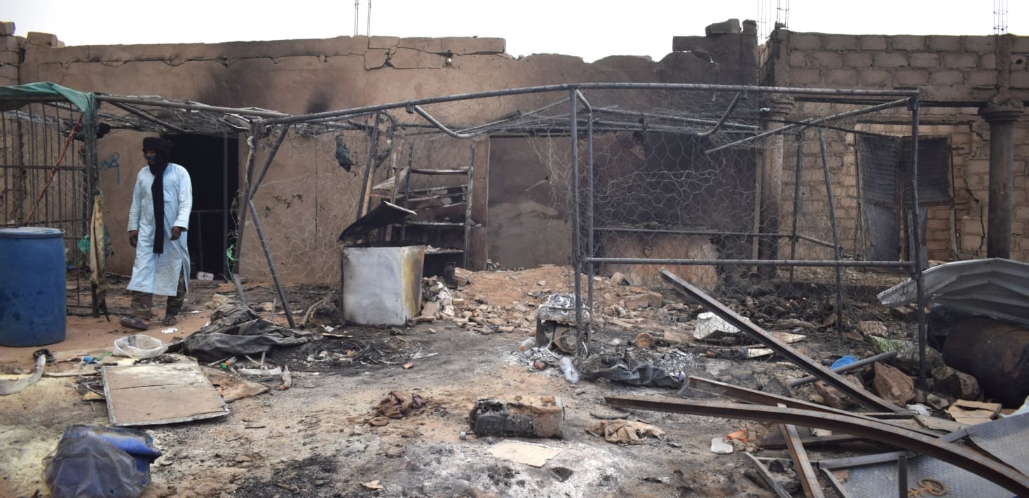 A man in a white robe and dark scarf walks through the burned out ruins of a building. The ground is littered with twisted charred metal and ash.