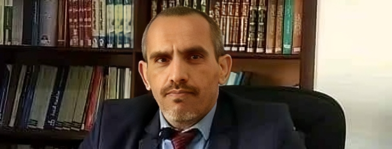 A man with a serious expression on his face wearing a suit and tie sits at a desk in an office chair in front of a bookshelf with many books.