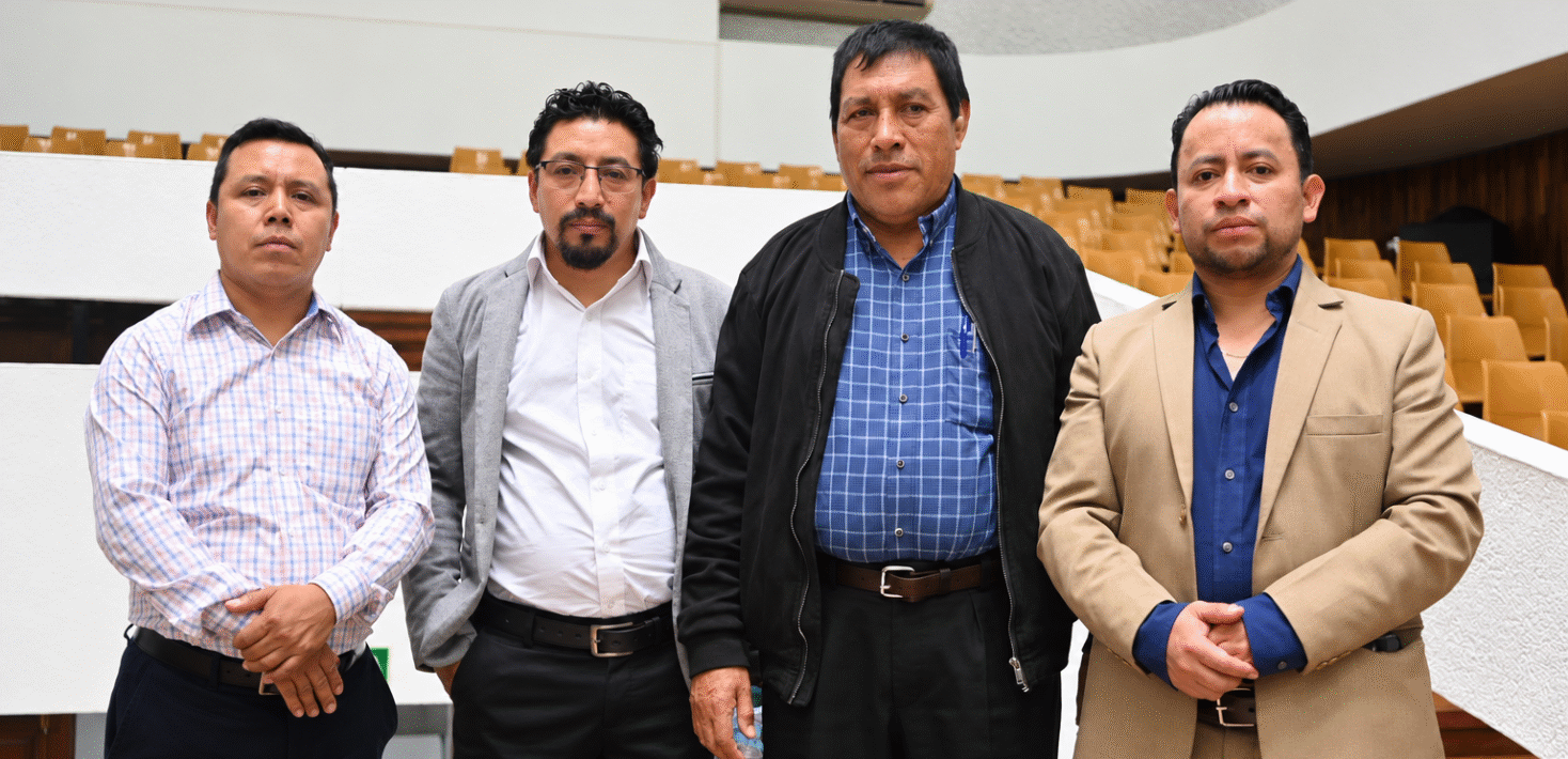 Four men stand in a line looking at the camera with solemn expressions in an empty lecture hall.