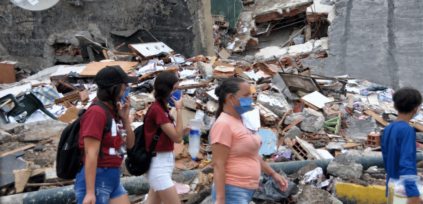 People wearing blue face masks walk by a pile of rubble.