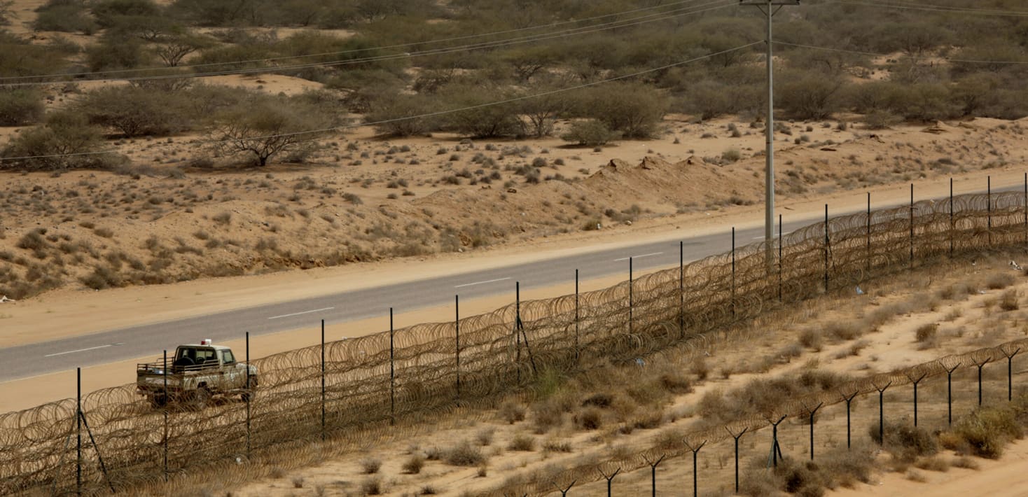A narrow paved road lined by a barbed-wire topped metal fence cuts through a desert landscape of sand and scrubby bushes. One truck sits parked in the sand by the side of the road.