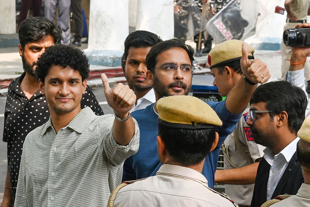 Two young men flash the thumbs up sign with confident expressions on their faces as uniformed police look on.