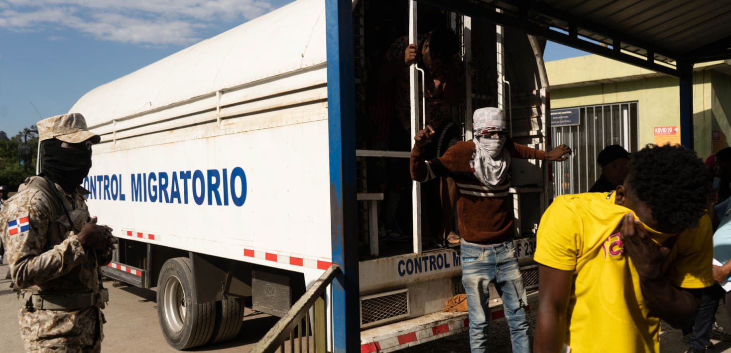 Black men with their faces covered by shirts jump out of the back of a parked truck as armed soldiers in Dominican uniforms watch.