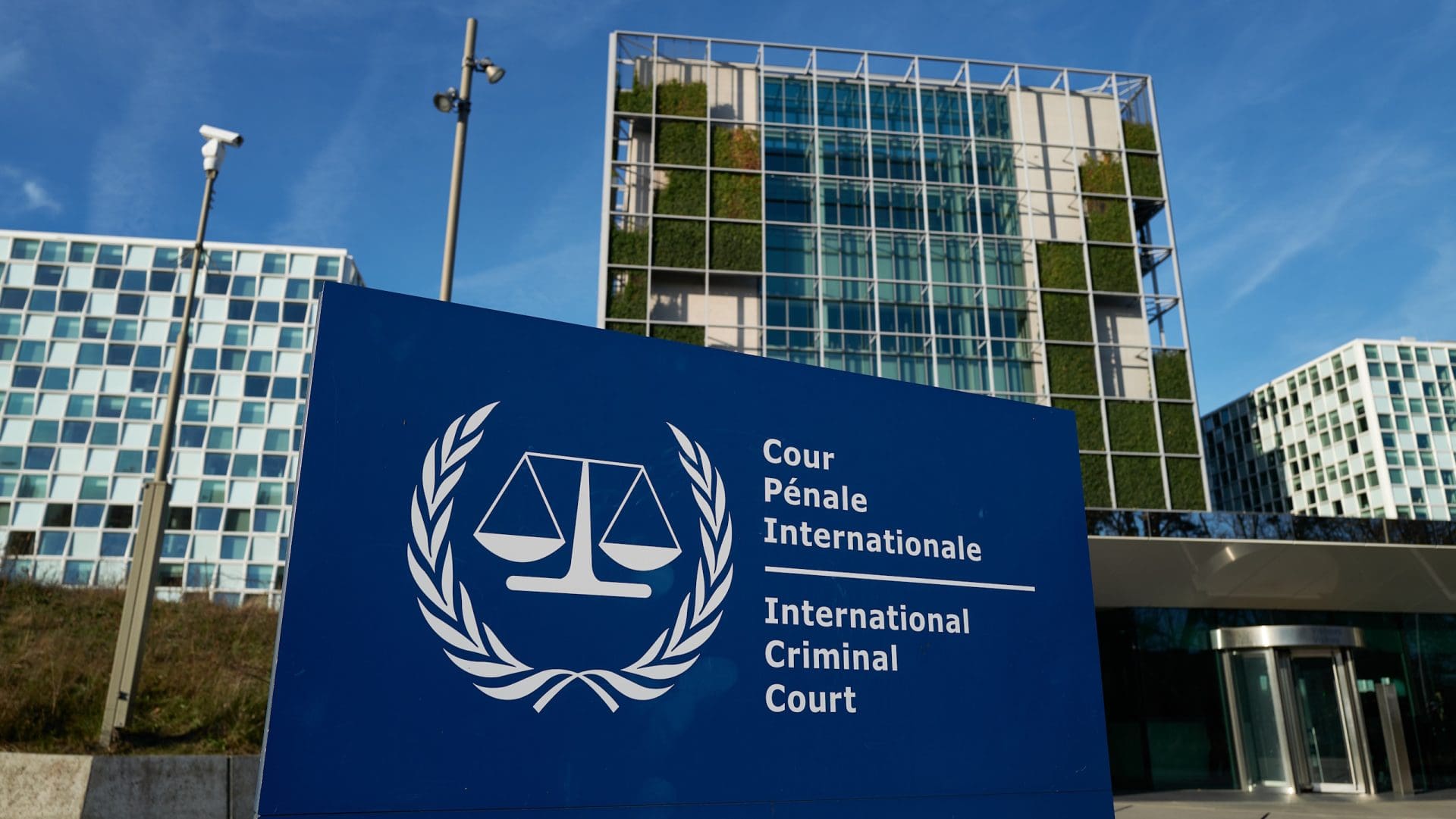 A blue and white sign that says International Criminal Court in English and French with the Court's logo, outside some office buildings. The sky is blue. It is daytime.