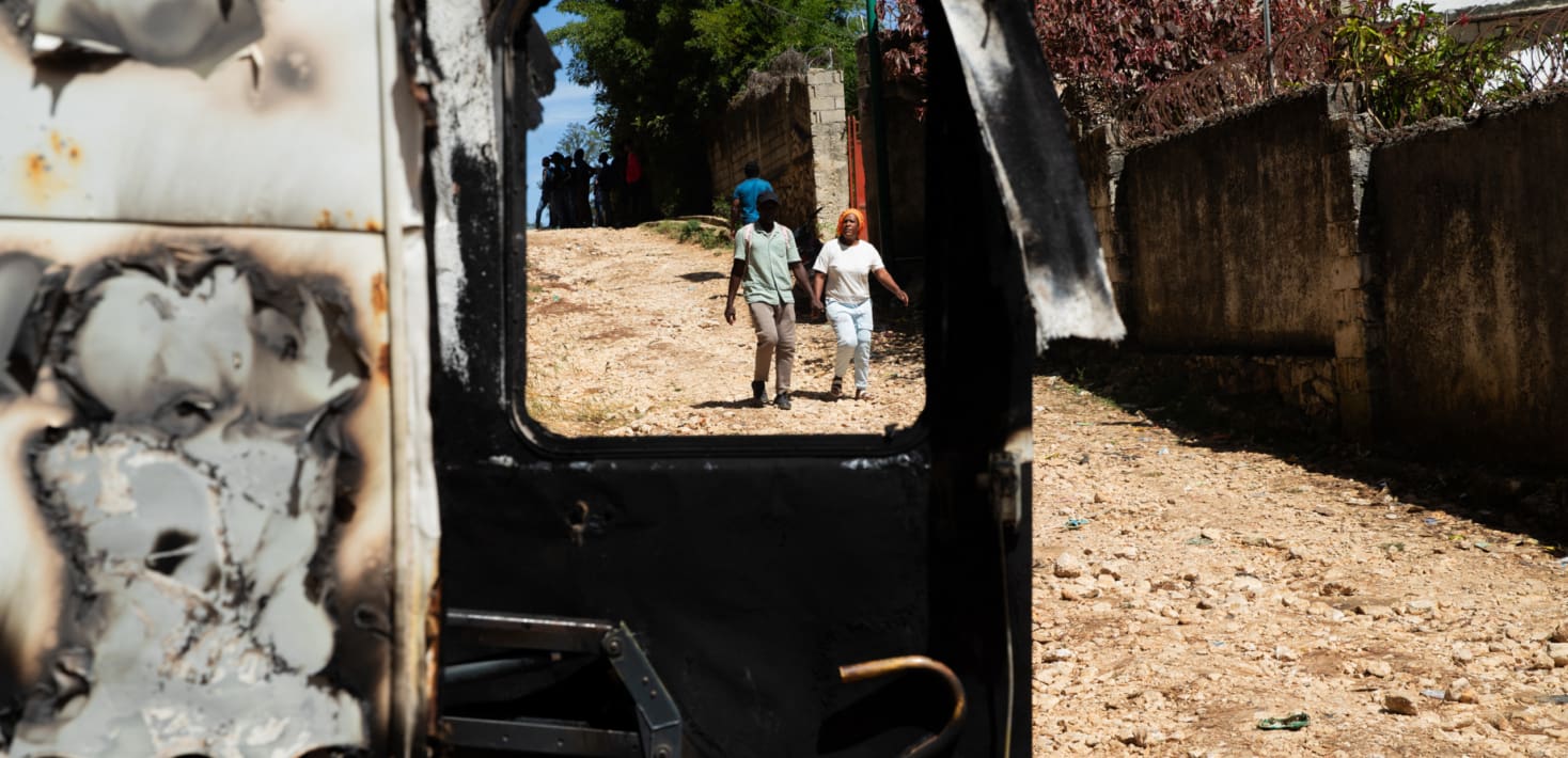 People walk on a dirt road next to a burned out car.
